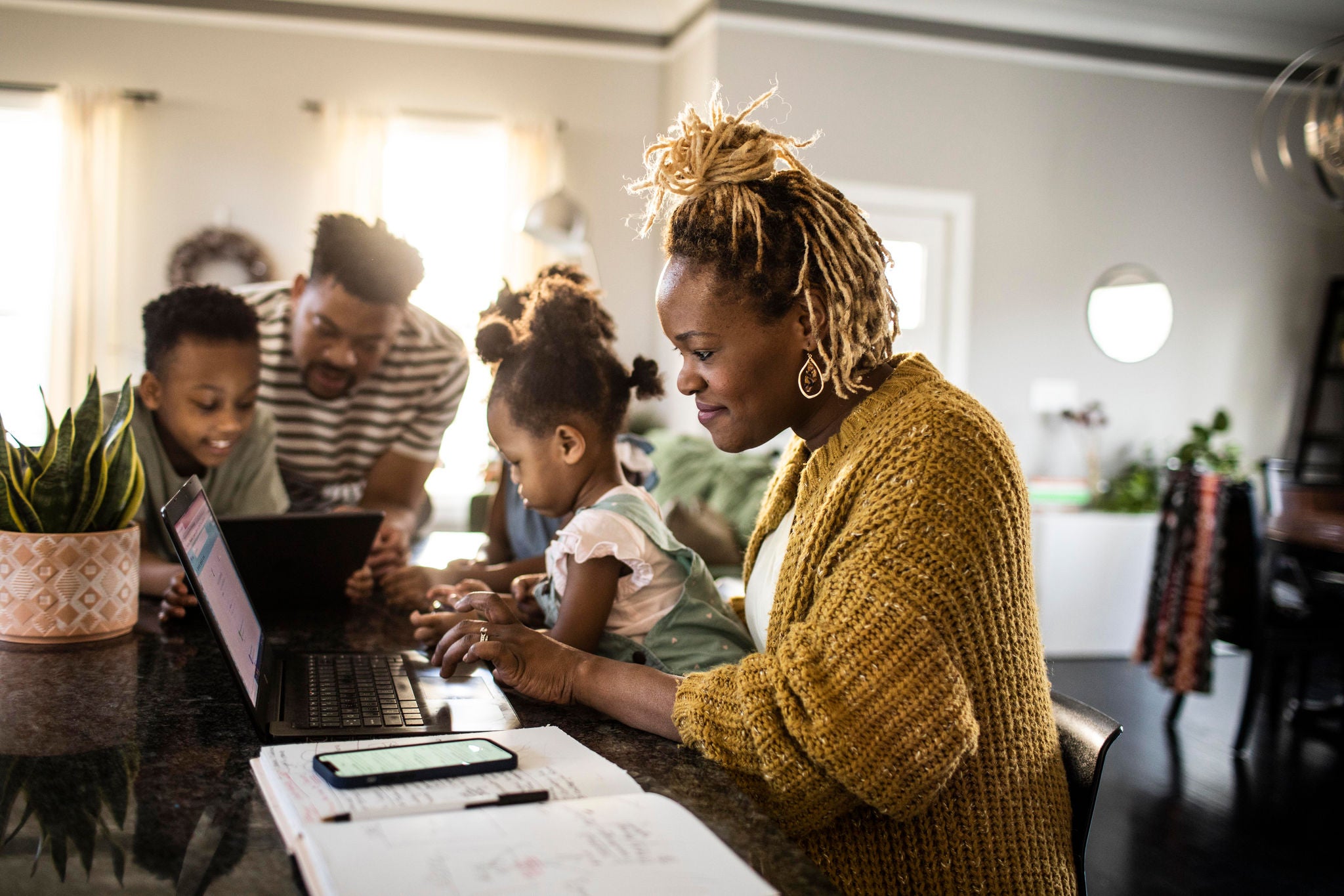 Mother working from home while holding toddler, family in background