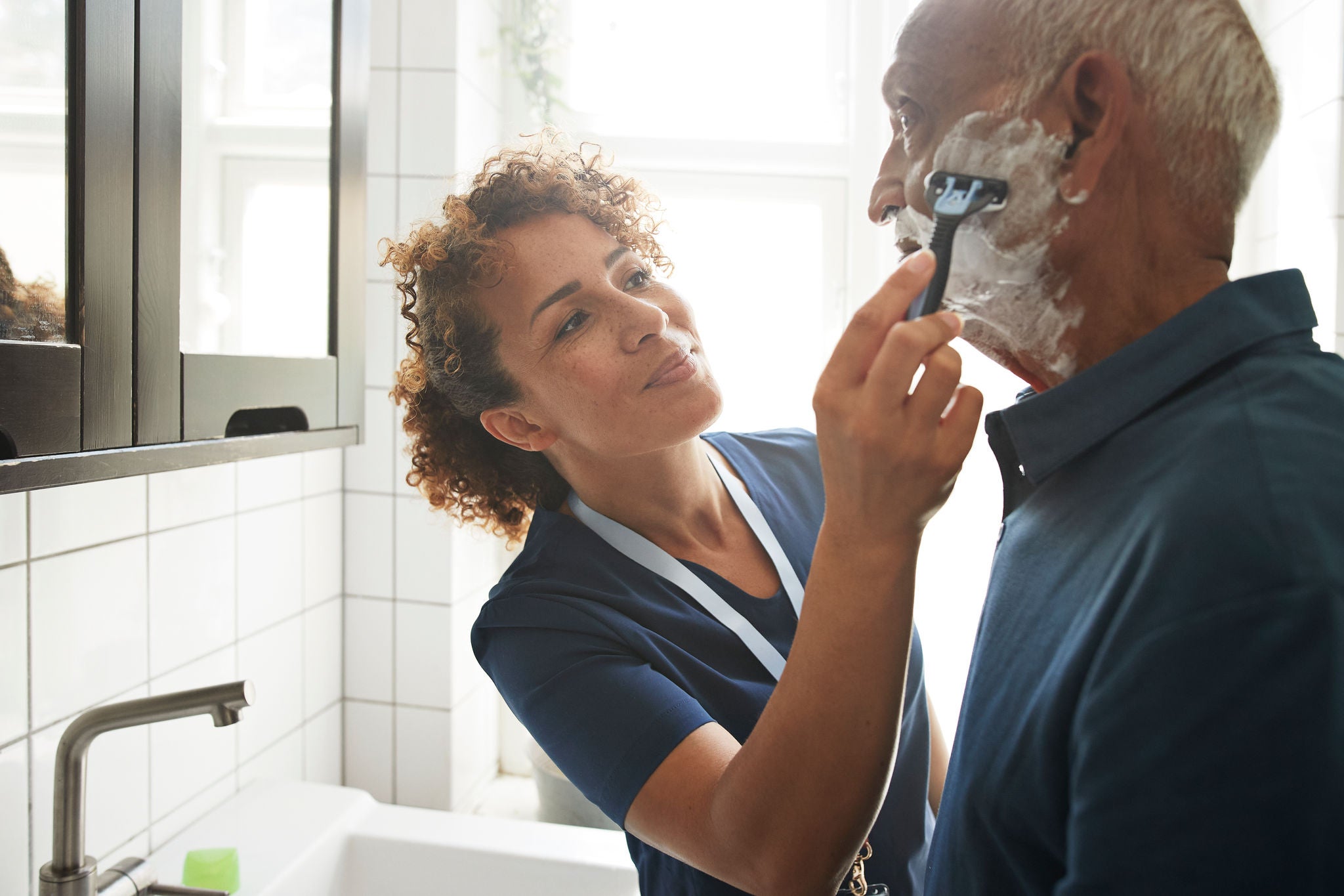 Female healthcare worker assisting senior man in shaving at bathroom