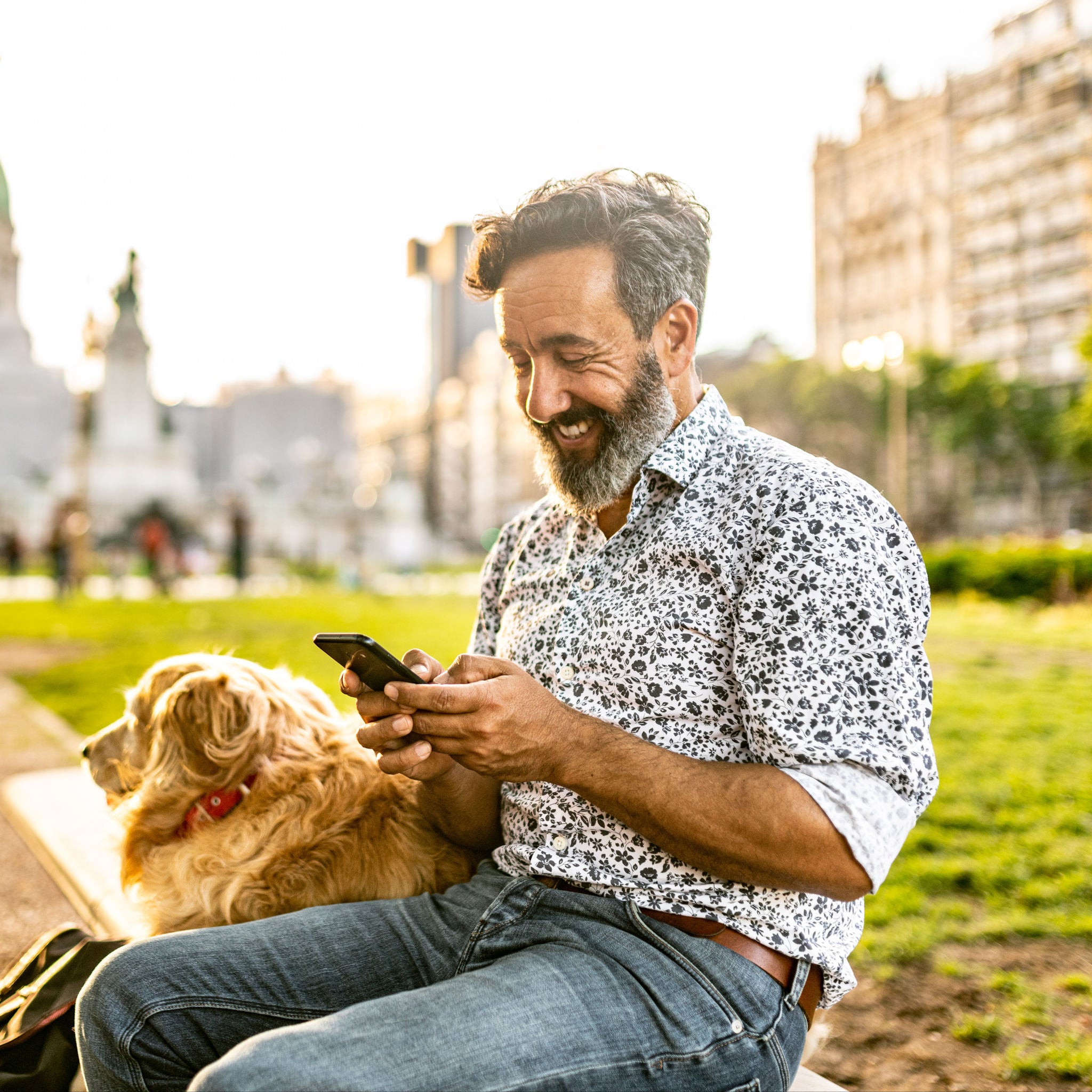 Mature man walking golden retriever uses mobile phone in public park