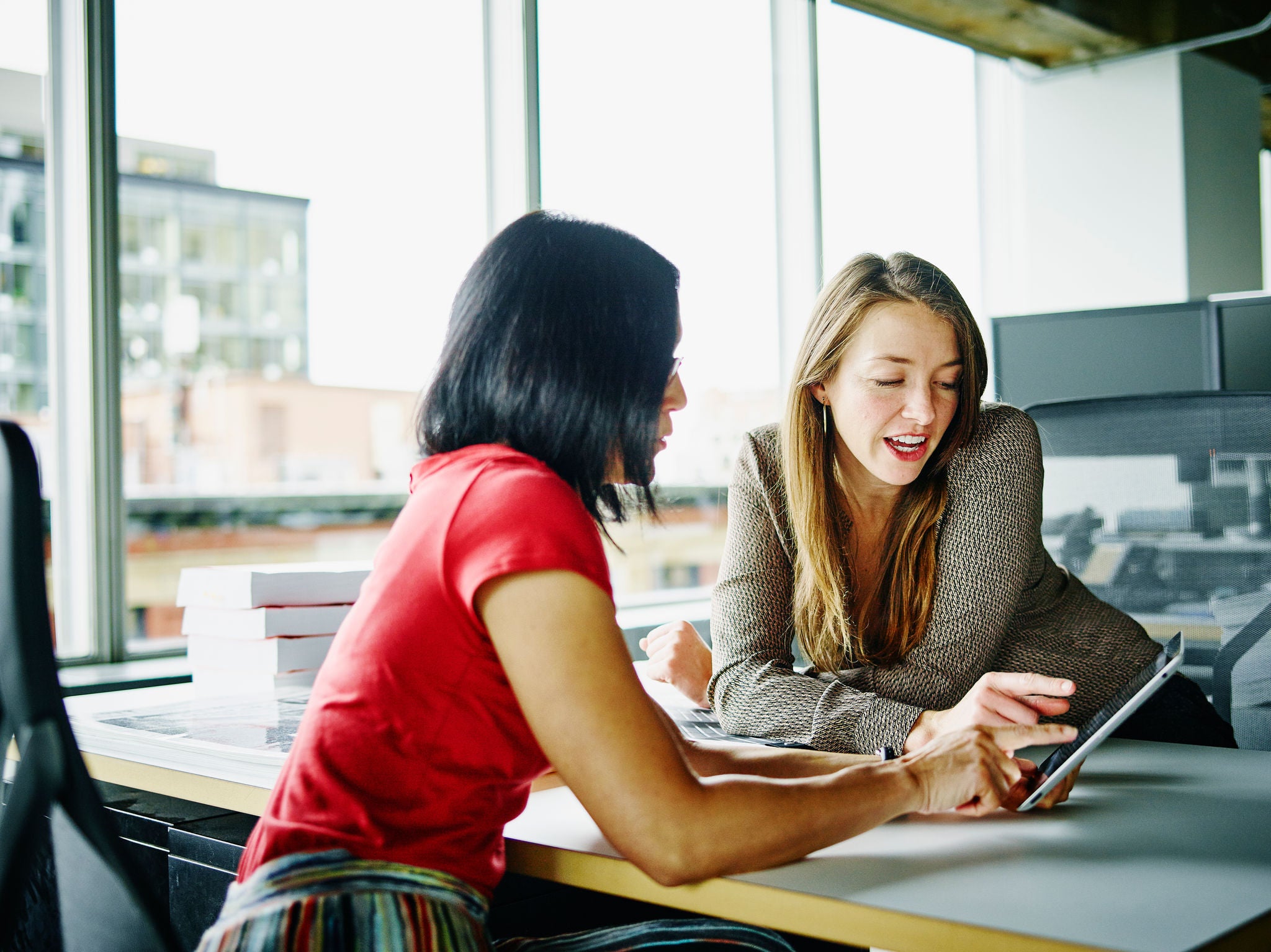 Two businesswomen sitting at workstation in office discussing project on digital tablet