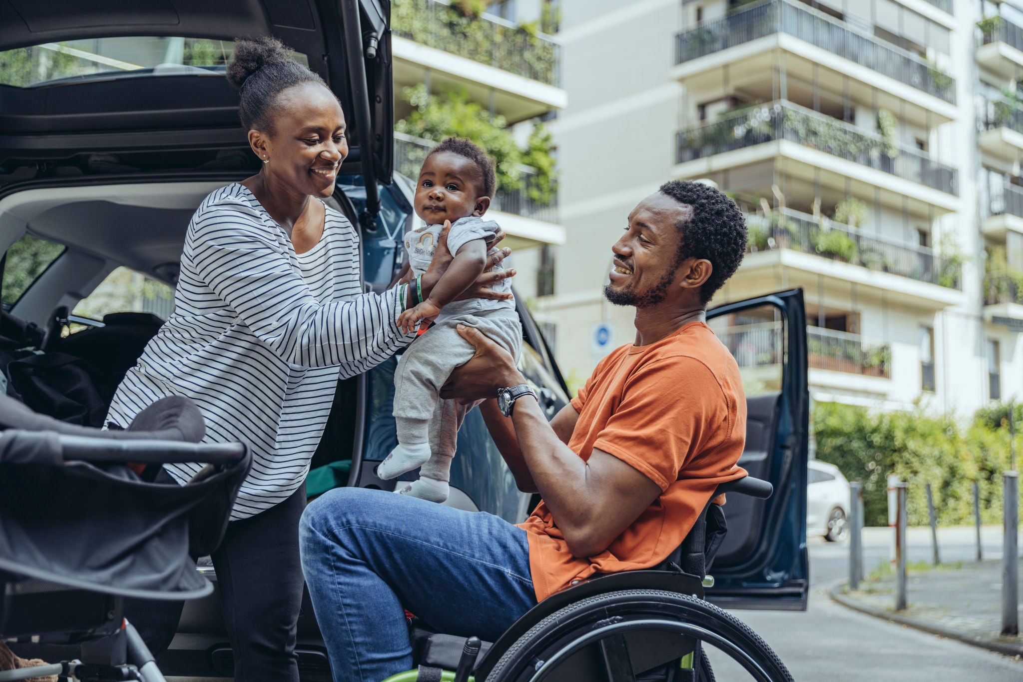 Mother giving son to father sitting in wheelchair by car