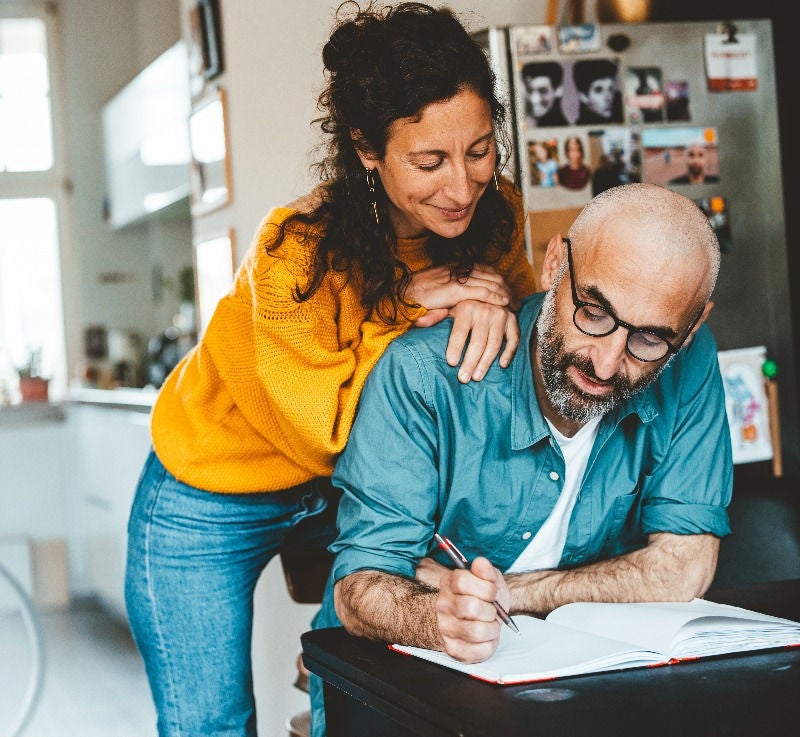 Woman leaning on man writing in notebook at home