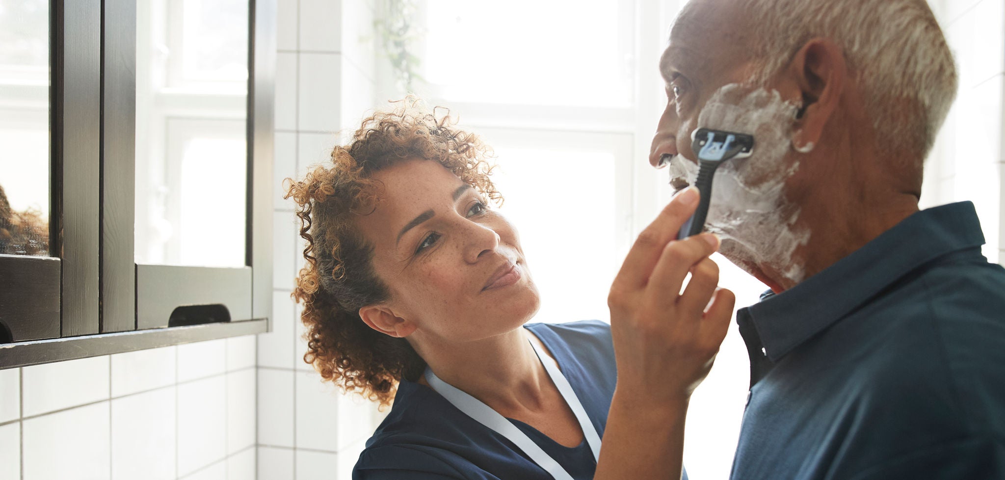 Female healthcare worker assisting senior man in shaving at bathroom
