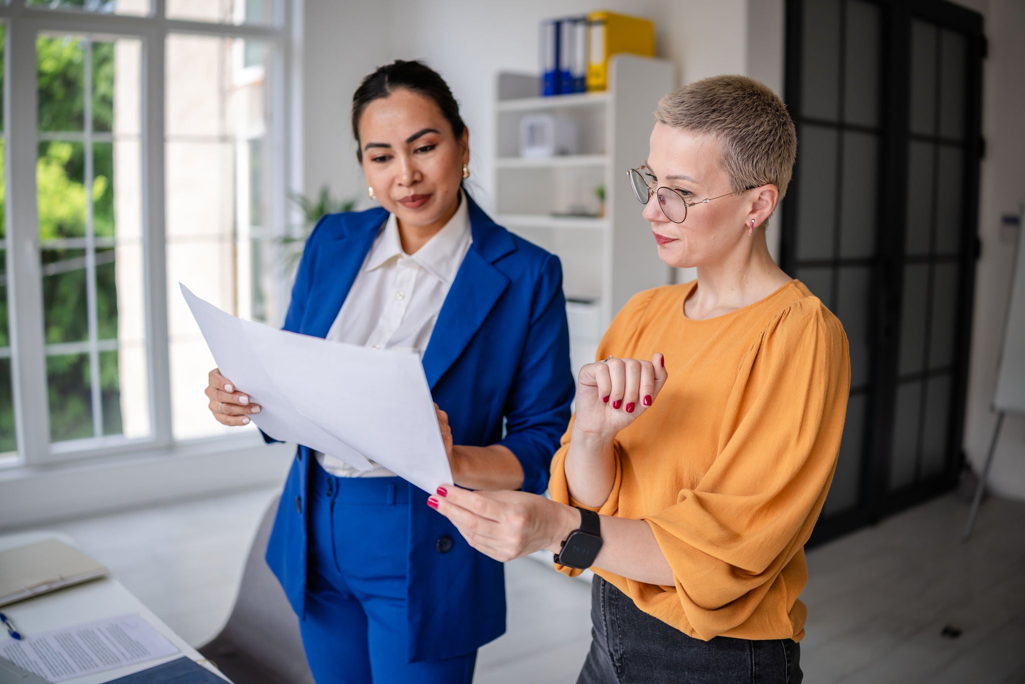 Inside a modern office, two women discuss and analyze documents. One is wearing a blue suit, while the other sports a yellow top. Natural light filters through large windows.